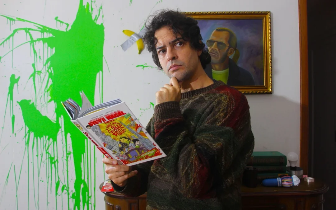 Juliano Enrico, com pose pensativa, segura um exemplar do novo livro do universo do Irmão do Jorel, que se chama "Steve Magal Contra o Aquecimento Global Brutal"