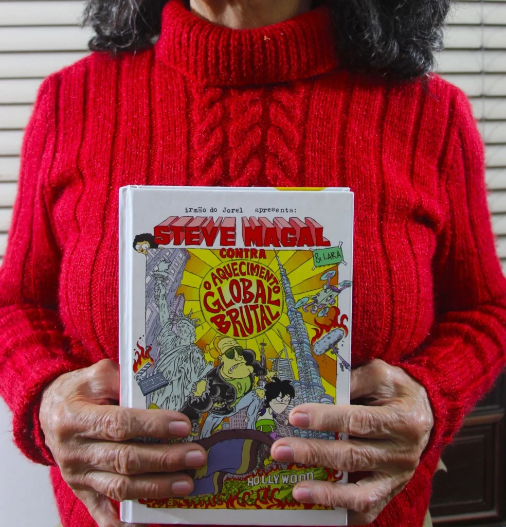 O novo livro do universo do Irmão do Jorel, protagonizado por Steve Magal, sendo segurado pela Dona Danuza da vida real
